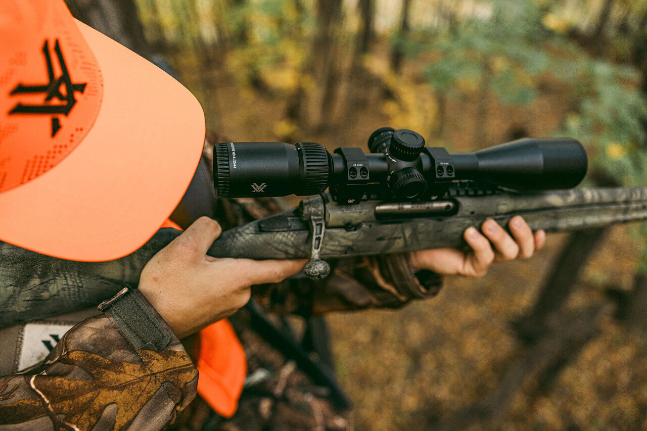 Eine Person in Camouflage-Kleidung und orangefarbener Kappe hält ein Gewehr mit Zielfernrohr in einem bewaldeten Gebiet.