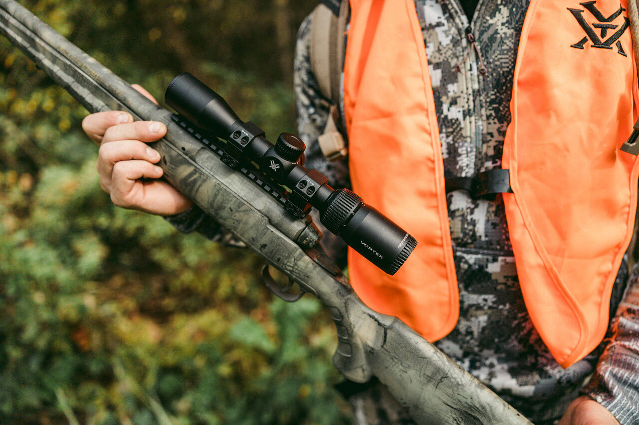 Ein Jäger hält eine Flinte mit einem montierten Visier in einem Wald, gekleidet in einer orangefarbenen Weste.