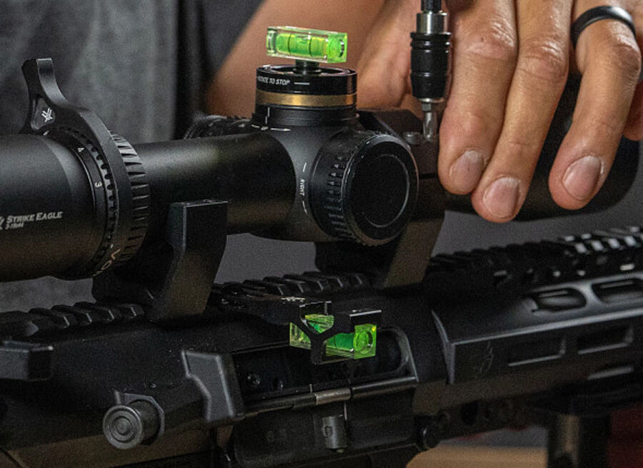 Nahaufnahme eines Zielfernrohrs mit integrierter Wasserwaage, das auf einem Gewehr montiert wird, während eine Hand Werkzeuge zur Justierung nutzt