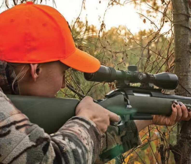 Jäger mit orangefarbener Mütze zielt mit Gewehr im Wald Person in Tarnkleidung und orangefarbener Mütze zielt mit Gewehr in einem bewaldeten Gebiet