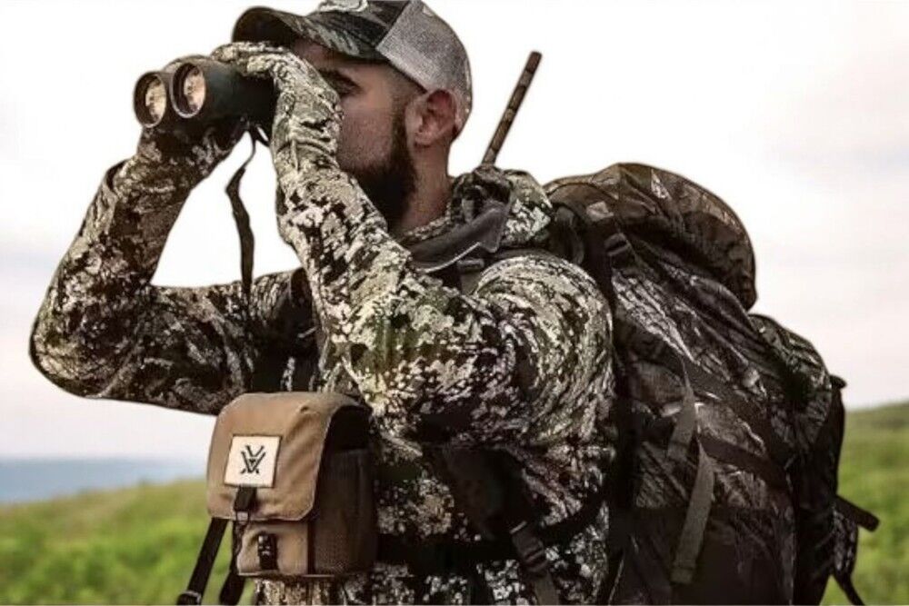 Jäger mit Tarnkleidung und Fernglas in der Natur Ein Mann in Tarnkleidung beobachtet mit einem Fernglas die Umgebung und trägt einen großen Rucksack.
