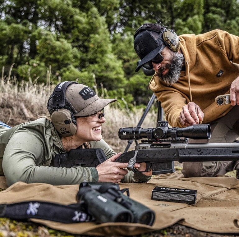 Outdoor-Schießtraining mit modernem Gewehrzielsystem Zwei Personen beim Schießtraining im Freien, mit Gehörschutz und moderner Zieloptik, auf einem Schießfeld mit Wald im Hintergrund.