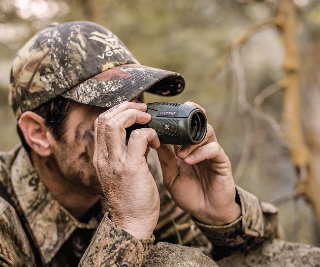 Ein Jäger in Tarnkleidung hält ein Fernglas und beobachtet die Umgebung im Wald.