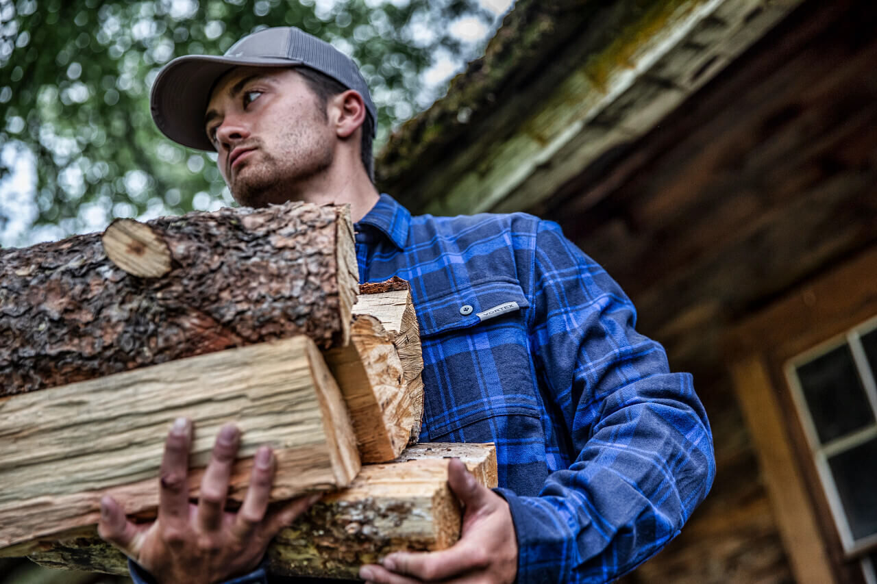 Ein Mann mit grauer Kappe und blauem Flanellhemd trägt mehrere Holzscheite vor einer Holzhütte