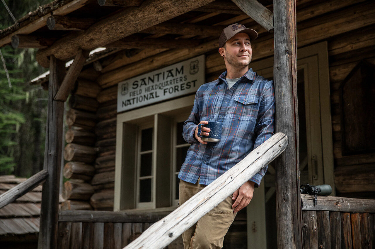 Ein Mann in einer karierter Hemdjacke steht auf der Veranda einer Blockhütte und hält eine Tasse in der Hand. Im Hintergrund ist ein Schild mit der Aufschrift 'Santiam Field Headquarters National Forest' zu sehen.