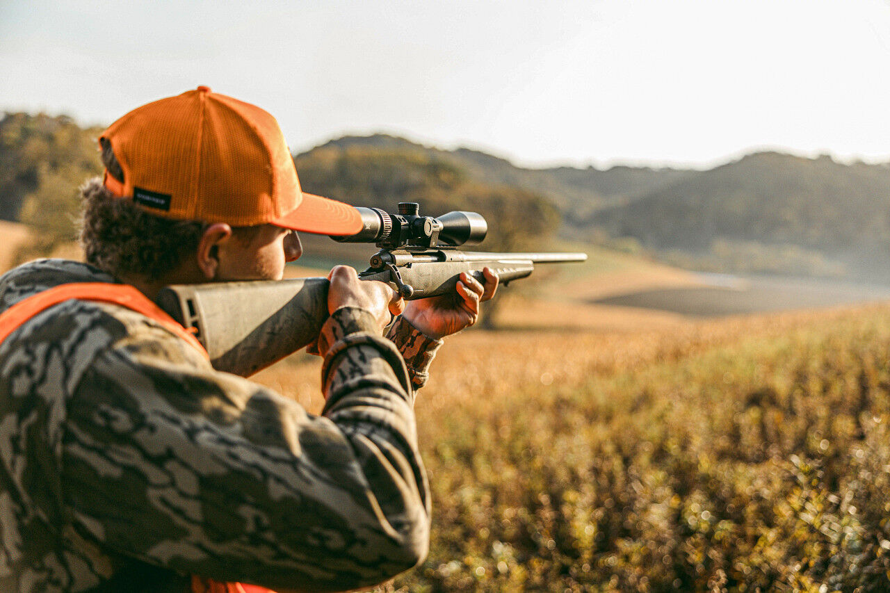 Ein Jäger in orangefarbener Mütze und Tarnkleidung zielt mit einem Gewehr auf einem Feld im Freien bei Sonnenschein.