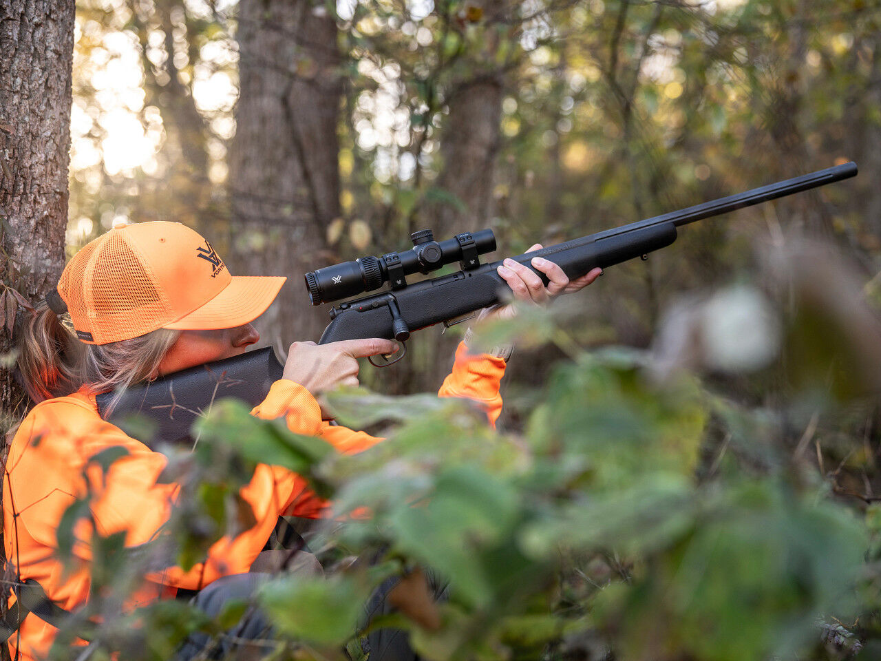 Eine Frau in orangefarbener Kleidung und Basecap zielt mit einem Gewehr im Wald