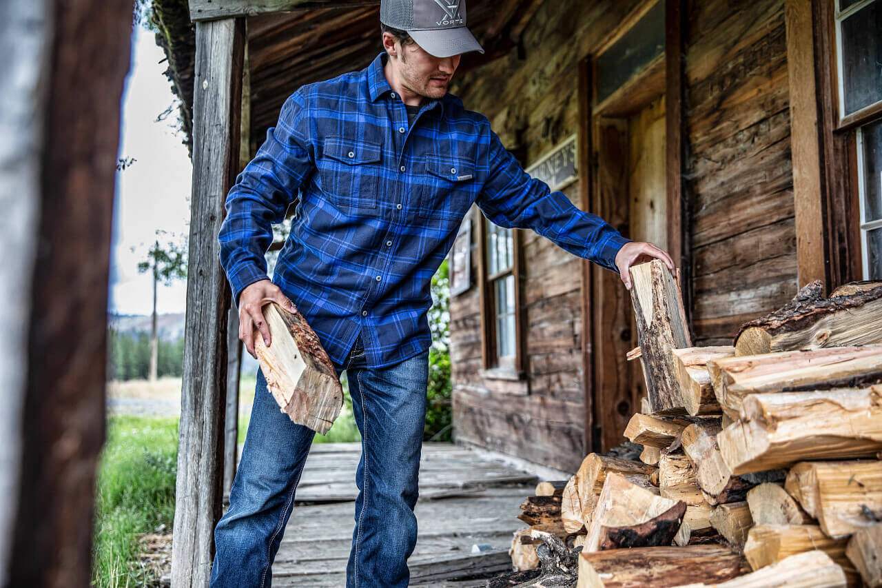 Eine Person in blauer kariertem Hemd stapelt Holzscheite auf einer Veranda eines alten Holzhauses.